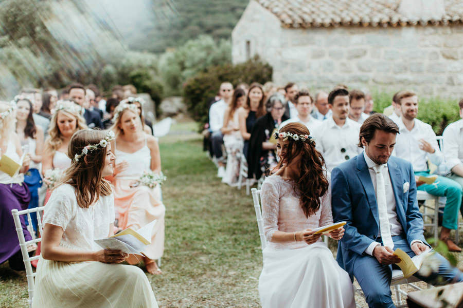 Guests seated outdoors at a countryside wedding in Sardinia, including a man in a blue suit and women wearing flower crowns.