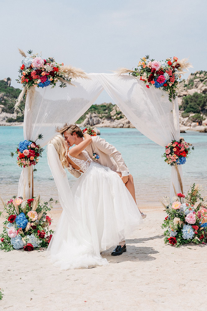 Sposi che si baciano sotto un arco floreale sulla spiaggia durante il loro matrimonio