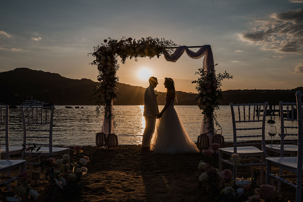 Cerimonia al tramonto allestita sulla spiaggia con arco floreale e vista sul mare